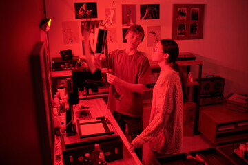 Teenage boy discussing developed photo prints and doing selections while working together in darkroom lit by red light young photographers in studio copy space