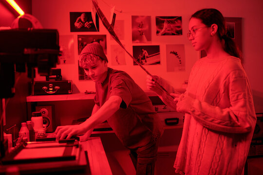 Two young photographers working together with analog photography busy with creative process in darkroom lit by red neon lights copy space