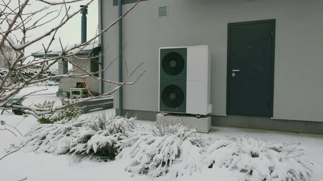 Heat pump unit outside a modern house surrounded by snow-covered plants, showcasing winter energy efficiency.