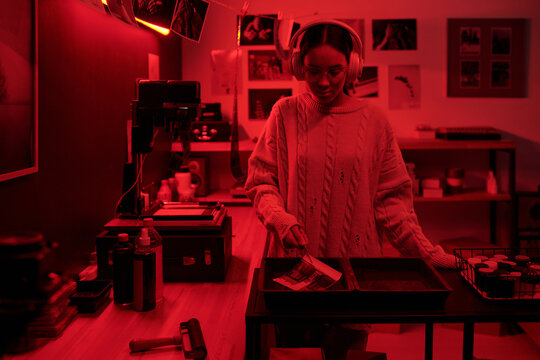 Waist up portrait of teenage girl wearing headphones developing analog photos in trays working in darkroom lit by red light photographer in studio copy space