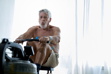 Staying fit and healthy. Senior shirtless man with an EKG mobile device exercising on rowing a machine at home