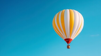 Fototapeta premium Yellow and white hot air balloon ascends against a clear blue sky. The balloon is the central focus, with a small wicker basket visible underneath