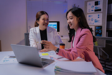 Two happy Asian businesswomen are discussing marketing strategy pointing at laptop in office at...