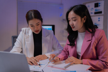 Two Asian businesswomen collaborating at night in an office, analyzing financial charts on a laptop to strategize business plans and market development