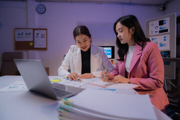 Two Asian businesswomen working late at night, collaborating on a project with a laptop while analyzing financial charts and graphs in a professional office