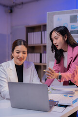 Obraz premium Two asian businesswomen are working together in the office at night, using a laptop and analyzing reports and statistics, discussing new strategies