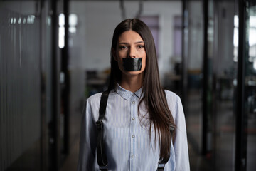 Serious woman standing in office with black tape over her mouth, symbolizing censorship,...