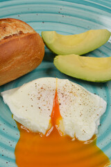 A close-up shot of a poached egg with a runny yolk, served with avocado slices and a bread roll on a light blue plate