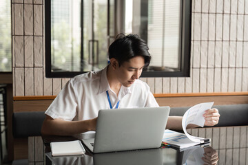 Young professional analyzing documents while working on laptop in modern office, focusing on data and project details.