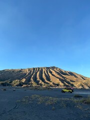 Serene panorama displays the beautiful Mount Bromo in Indonesia, showcased under a bright blue sky, captured in a wide shot alongside a passing jeep.