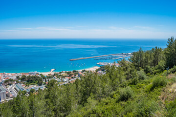 Fototapeta premium Green forest of the Arrábida natural park with construction of the village of Sesimbra next to the beach and marina in the background, PORTUGAL