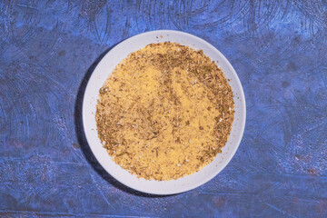A white ceramic plate filled with a mixture of yellow seasoning and dried herbs, placed on a blue surface.