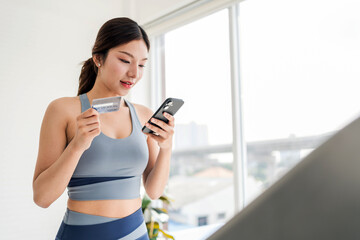 Young Asian woman in active wear shops online using her smartphone and credit card after a workout....