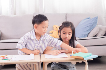 Asian young boy and girl studying together at home. Boy assisting girl with homework, pointing at paper. Cozy living room setting with sofa and stuffed toy in background. Education teamwork.