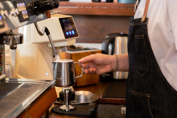  Barista frothing milk with steam wand standing in bar Stainless steel pitcher under steam wand with foam forming, Close-up of barista hand, Professional coffee preparation in  small business cafe