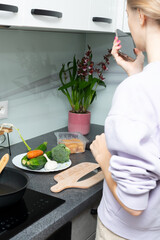 Young woman in the kitchen uses AI technology to help prepare a delicious and healthy lunch of vegetables and chicken fillet. Modern technology, lifestyle