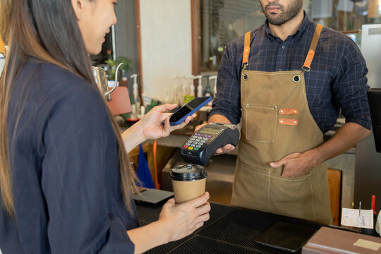 Young Indian baristas or cashier workers man holding a payment machine for scan, cashless technology and credit card payment concept. Customer using smartphone for payment to owner at cafe restaurant