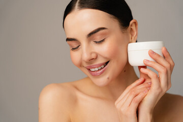 The image depicts a person smiling while holding a jar of cream close to their face, with a focus...
