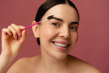 Fototapeta premium Cheerful individual using a cosmetic tool to enhance eyebrow appearance, standing against a pink background, portraying beauty and self-care.