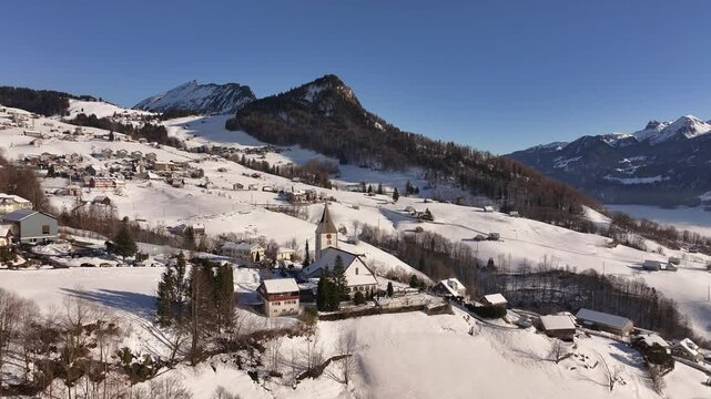 Scenic aerial view of Amden, Switzerland covered in fresh snow, stunning mountain backdrop, charming village houses and picturesque church.