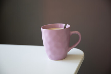 Close up of a pastel pink cup with hot coffee drink on white table, dark grey wall background 