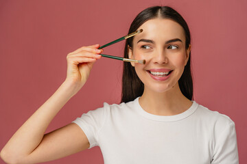 Obraz premium Smiling individual playfully holding makeup brushes in a creative pose, set against a vibrant pink backdrop that highlights cheerful and engaging beauty concepts.