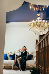 A woman sits on an elegant sofa with carved wooden elements and light-colored upholstery. There are blue pillows on the sofa. The woman is wearing a long green dress and black tights