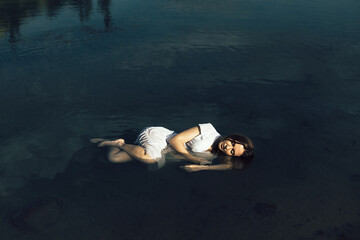 A woman in a white dress lies on the surface of the water, curled up in a fetal position. The water is clear and the bottom is visible. Her eyes are closed and she looks relaxed.