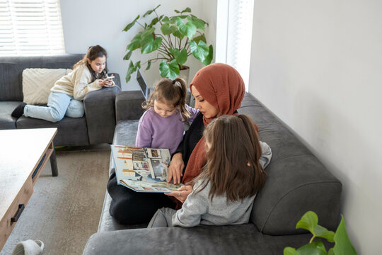 A woman in a hijab reads a book to two young girls on a sofa, while another girl uses a smartphone on an armchair in a cozy living room, Netherlands