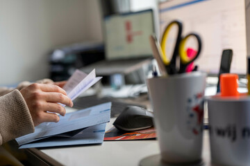 A person sorts through mail at a cluttered desk, featuring a computer monitor, keyboard, mouse, documents, and a pen holder with scissors, Nijmegen, Netherlands