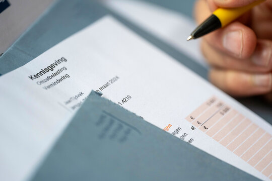 A hand holding a yellow pen is poised over a document, partially obscured by a blue envelope, Nijmegen, Netherlands