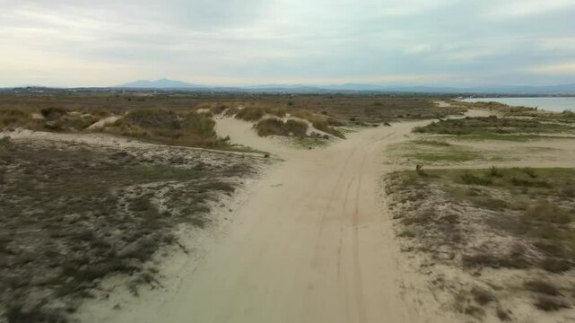 Day view low flight over an empty dirt road and an expansive desert landscape with sandy paths next to the sea under a cloudy sky in Greece.
