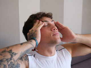Portrait of a young tattooed man in a white sleeveless t-shirt sitting on the couch touching his...