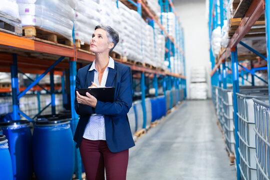 Professional woman inspecting inventory with clipboard in large warehouse aisle