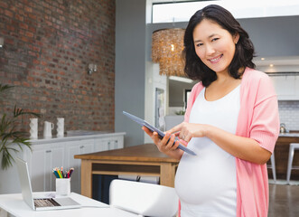 Pregnant Asian woman using tablet and smiling at home in modern kitchen, copy space