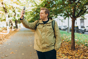 Obraz premium Smiling 30s man with backpack taking photo on park background. Selfie time. Holidays, rest, blogging banner. Charming male wearing stylish outfit have fine