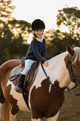 A young girl rider in a helmet and equestrian boots sits on her beloved horse at the stable,...