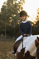 A young girl rider in a helmet and equestrian boots sits on her beloved horse at the stable,...
