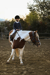 A young girl rider in a helmet and equestrian boots sits on her beloved horse at the stable, enjoying a peaceful horseback ride.