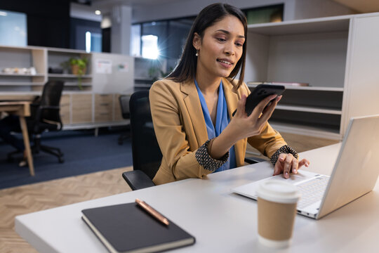Businesswoman using smartphone and laptop at office desk, multitasking efficiently