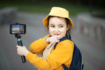 Cute little girl taking selfie with mobile phone looking at camera in a park. Kid blogger recording video vlog.