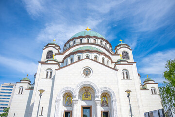 Saint Sava Cathedral stands majestically under a clear blue sky. The sunlight illuminates its intricate architecture, while surrounding greenery adds to the serene summer atmosphere.