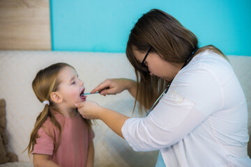 Female pediatrician in white coat with stethoscope examining throat of sick little girl with tongue depressor during home visit, providing individual medical care. Doctor's appointment at home.