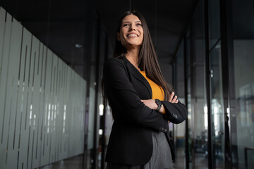 Confident young woman, entrepreneur, or manager standing in a modern office hallway, smiling with arms crossed. Concept of leadership, career success, and professional confidence