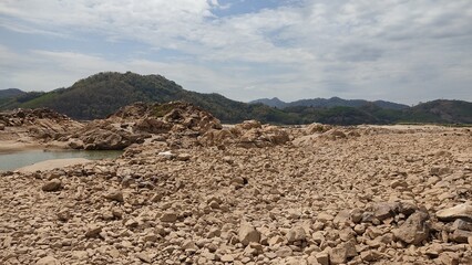Rocky dry riverbed landscape with scattered stones and boulders under a partly cloudy sky. Mountainous background with green hills, depicting drought, climate change, and natural erosion effects
