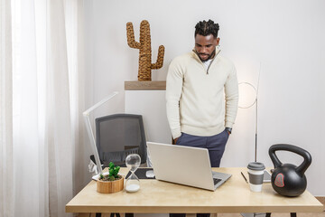 Afro-American man working remotely at home in a stylish, modern workspace with a laptop and...