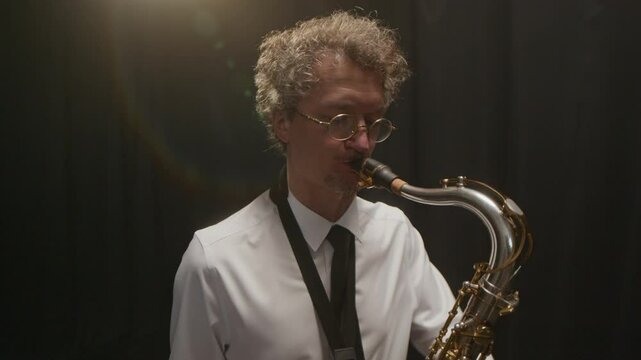 Tilt up shot of skilled middle-aged Caucasian male musician wearing classic outfit performing bebop on saxophone, playing masterfully on dark background