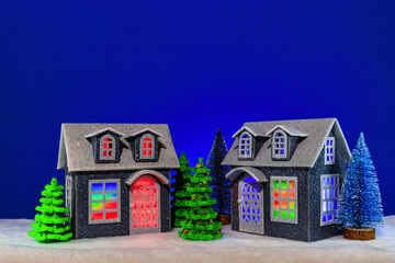 Two cozy houses with bright light in the windows surrounded by Christmas trees on a blue background