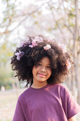 A young woman with voluminous curly hair adorned with pink blossoms smiles warmly. Dressed in a...