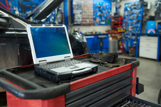 Rugged laptop computer displaying diagnostic software sits on a red toolbox in a busy auto repair shop, with a pair of mechanic's gloves nearby, suggesting ongoing vehicle maintenance